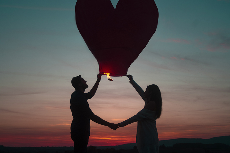 Pareja de enamorados sosteniendo un farolillo volador con forma de corazón en una puesta de sol