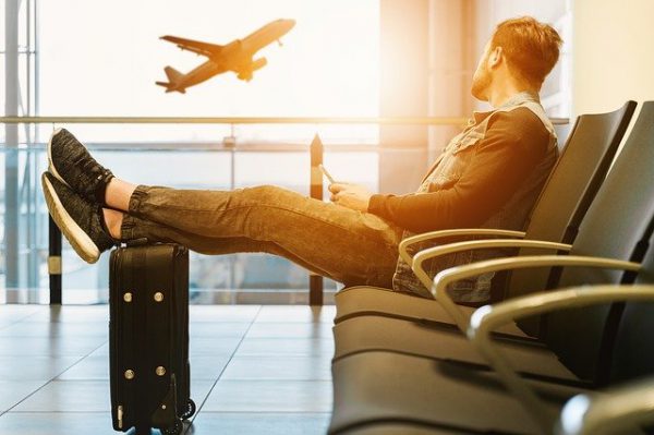 Hombre sentado en los asientos de un Aeropuerto con los pies apoyados sobre su maleta y viendo despegar un avión